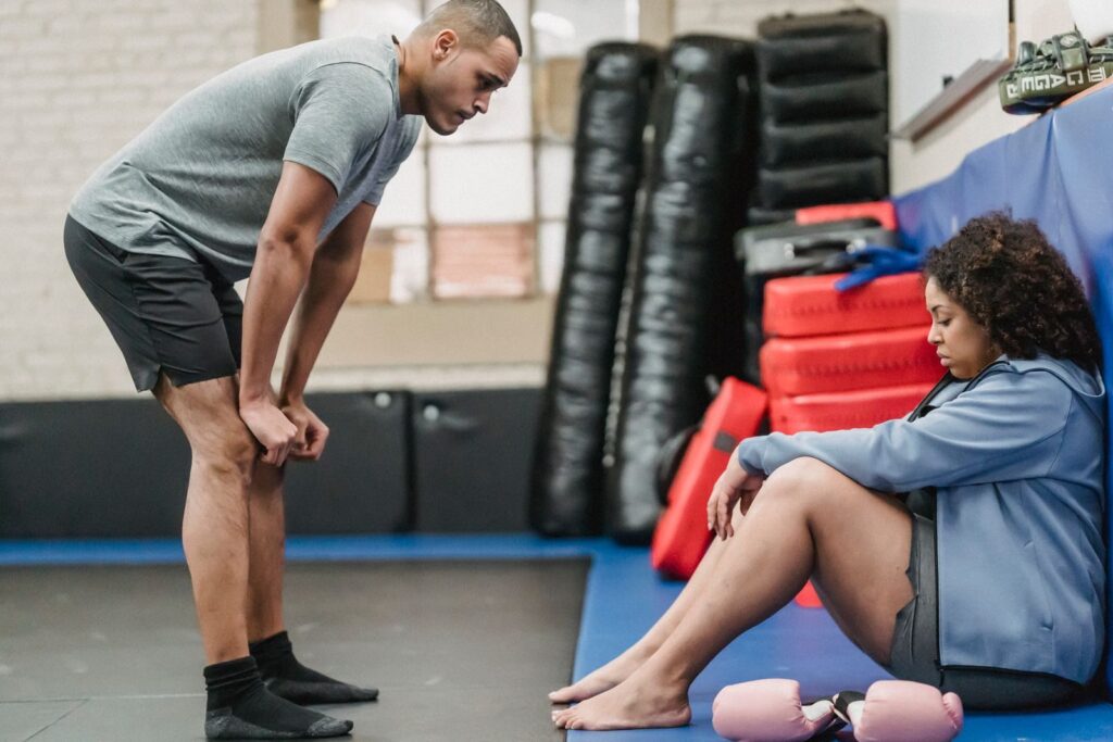 Full body side view of muscular personal coach standing near tired plump African American female during workout in gym with equipment